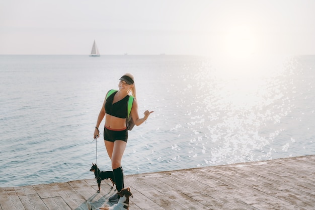 The young beautiful athletic girl with long blond hair in black clothes and two of her dogs doing sports at sunrise by the sea