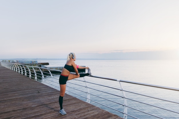 The young beautiful athletic girl with long blond hair in headphones listening to music and doing stretching at sunrise over the sea