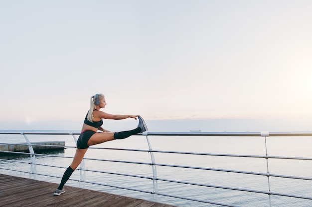 The young beautiful athletic girl with long blond hair in headphones listening to music and doing stretching at sunrise over the sea