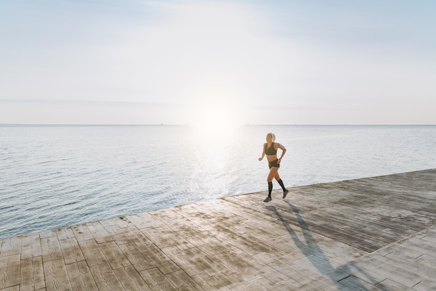 The young beautiful athletic girl with long blond hair in headphones listening to music and running at sunrise over the sea