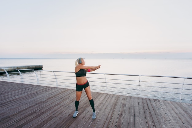 The young beautiful athletic girl with long blond hair in headphones listening to music and training at sunrise over the sea