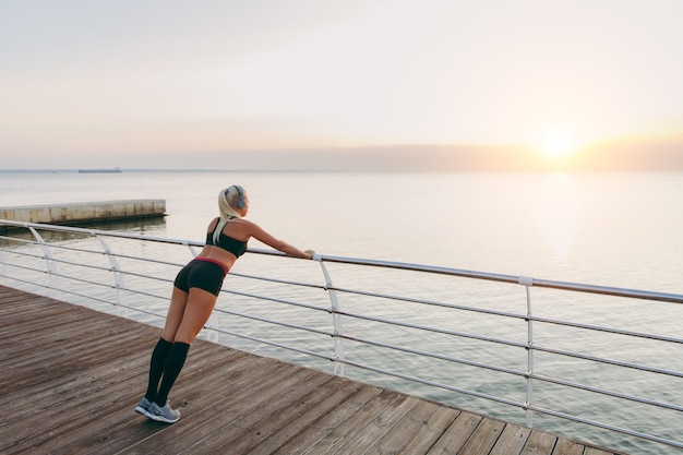 The young beautiful athletic girl with long blond hair in headphones listening to music and training at sunrise over the sea