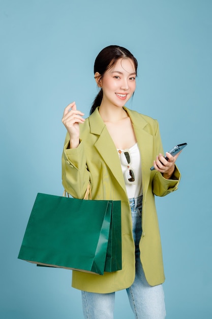 Young beautiful attractive Asian woman in casual fashion jacket and sunglasses holding shopping bags using mobile phone isolated on pastel blue background Female Consumerism lifestyle concept