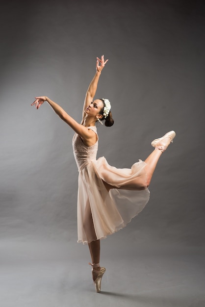 Young beautiful ballet dancer in beige swimsuit posing on pointes on light grey studio background