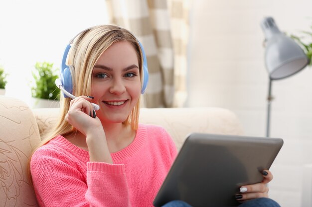 Young beautiful blond woman sit on the sofa in livingroom hold tablet in arms listen to music sunny