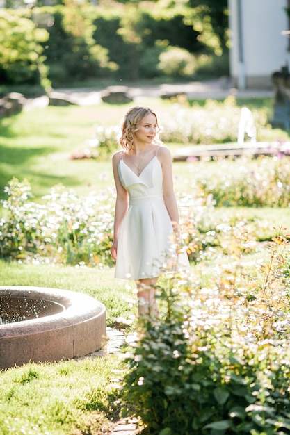 Young beautiful blonde 20s with curly hair in a white dress in the sunny sunset rays in the park A stylish model walks through the city's blooming park in summer