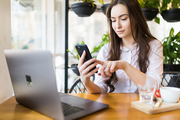 Young beautiful blonde female holding mobile phone while sitting with portable net-book in coffee shop interior, charming dreamy woman using cell telephone and laptop computer during rest in cafe