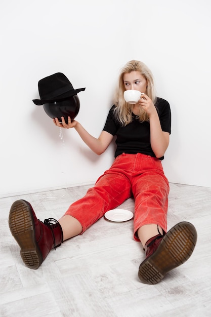 Young beautiful blonde girl holding black baloon in hat sitting on floor drinking coffee over white wall