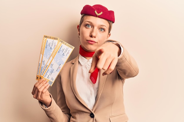 Young beautiful blonde girl wearing stewardess uniform holding boarding pass pointing with finger to the camera and to you confident gesture looking serious