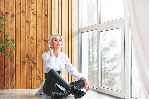 Young beautiful blonde model girl posing in a white shirt at home by the window