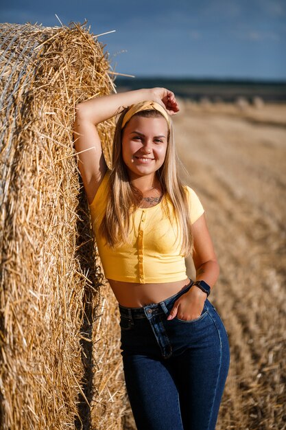 Photo a young beautiful blonde stands on a mown wheat field near a huge sheaf of hay, enjoying nature. nature in the village