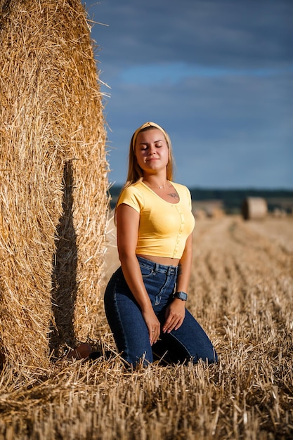 A young beautiful blonde stands on a mown wheat field near a huge sheaf of hay, enjoying nature. Nature in the village