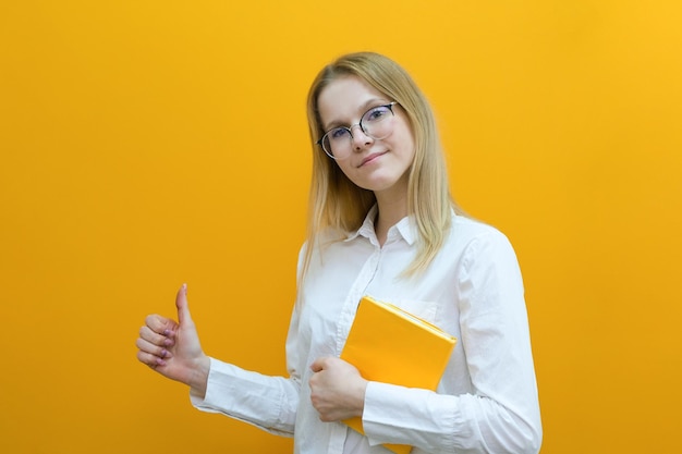 Young beautiful blonde student girl in glasses holds books on an yellow background