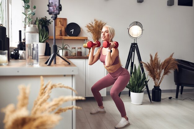 Young beautiful blonde takes care of her body while at home during quarantine. She trains people online through her mobile phone.