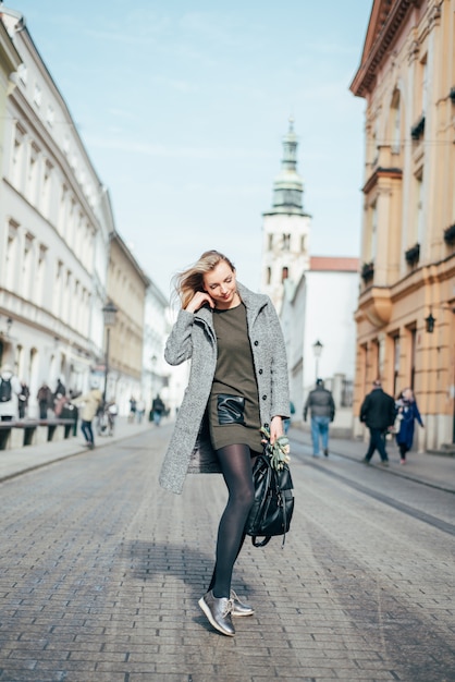 Young Beautiful  blonde woman in grey coat, dark green short dress and black tights walking in the city streets.