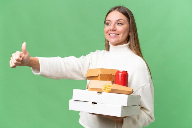 Young beautiful blonde woman holding takeaway food over isolated background giving a thumbs up gesture