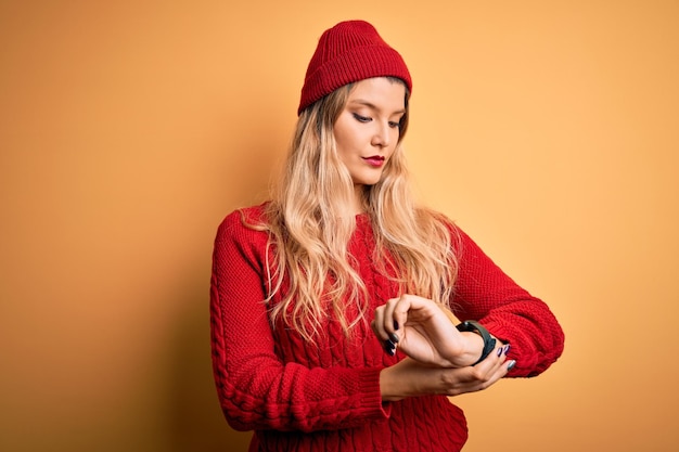 Photo young beautiful blonde woman wearing casual sweater and wool cap over white background checking the time on wrist watch relaxed and confident