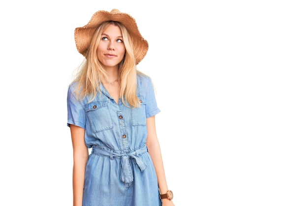 Young beautiful blonde woman wearing summer hat and dress smiling looking to the side and staring away thinking