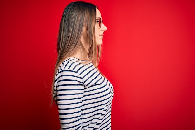 Young beautiful blonde woman with blue eyes wearing glasses standing over red background looking to side relax profile pose with natural face and confident smile