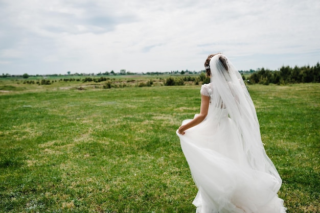 Young beautiful bride in an elegant dress is standing back on the field near the forest and holding bouquet flowers at nature Outdoors After the wedding ceremony