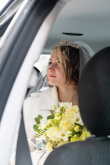 A young beautiful bride with a large bouquet of white roses sits in a car and looks away