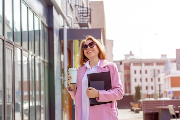 A young beautiful brunette businesswoman in sunglasses and a pink coat is walking along a city street in her hands she has a glass of coffee and a tablet a notebook