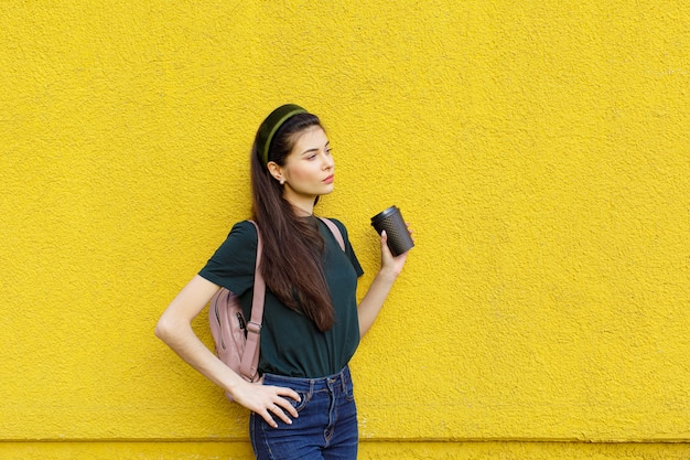 Young beautiful brunette girl in jeans a green T-shirt hair bands and a pink backpack posing on a yellow concrete wall