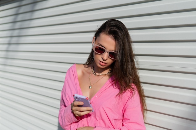 Young beautiful brunette in a pink shirt neck jewelry necklace trendy sunglasses on the background of a light garage fence looks at the phone screen