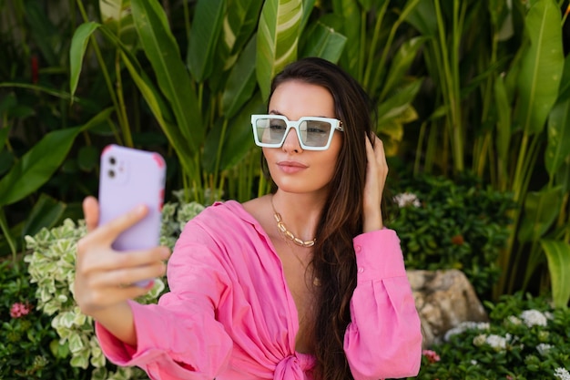 Young beautiful brunette in a pink shirt neck jewelry necklace trendy sunglasses sits on a background of tropical leaves takes a selfie on the phone