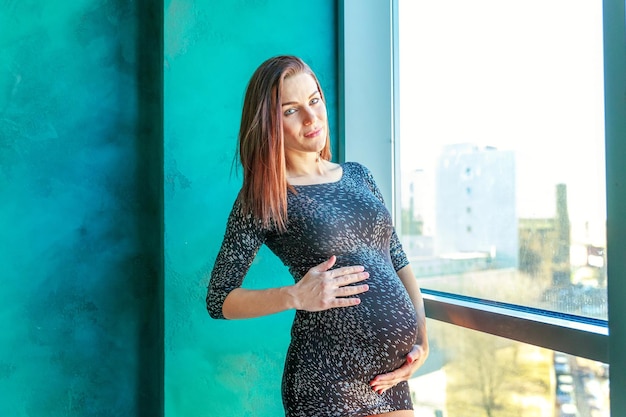 Young beautiful brunette pregnant woman standing near blue wall by window at home maternity pregnanc