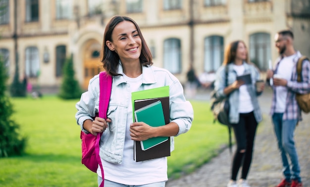 Young beautiful brunette student girl in denim clothes with backpack and books in hands is standing on a group of her student friends