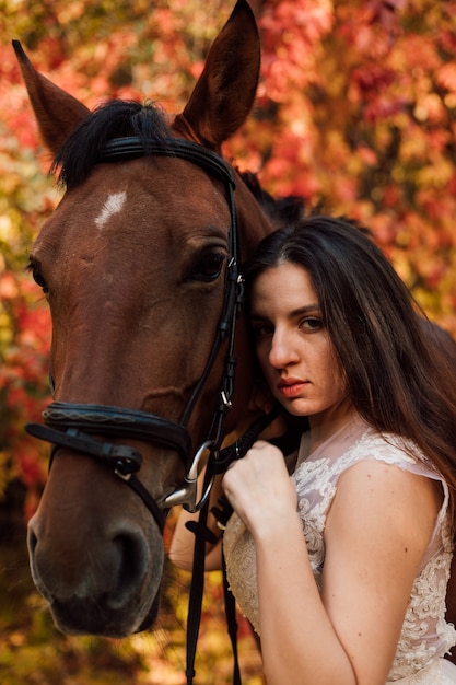 Young beautiful brunette in a white dress hugs a brown horse