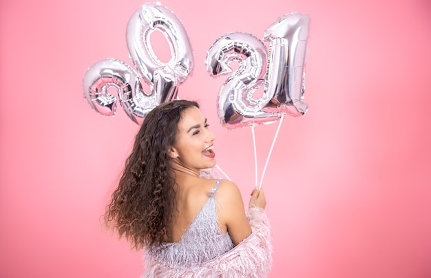 Young beautiful brunette with curly hair and an open back in profile posing on a pink wall with silver balloons for the new year concept