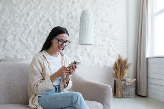 A young beautiful brunette woman in glasses is happy and smiling sitting on the sofa using the phone