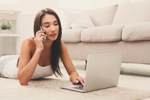 Young beautiful brunette woman relaxing at home while lying on floor carpet on her stomach talking on smarthone and using laptop. Technology and enjoyment concept.