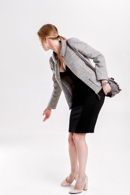 Young beautiful business woman blonde in black dress, jacket and purse looked back on gray background