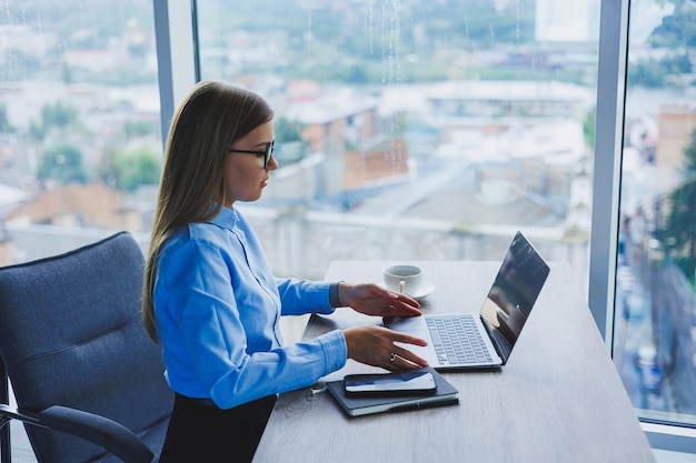 Young beautiful business woman in a blue shirt sitting in the office with a laptop and doing business