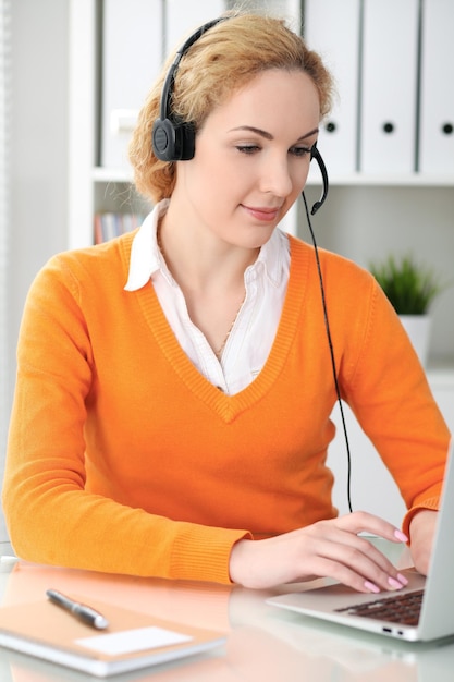 Young beautiful business woman in headset or call operator communicates by laptop computer Orange sweater is uniform of a girl