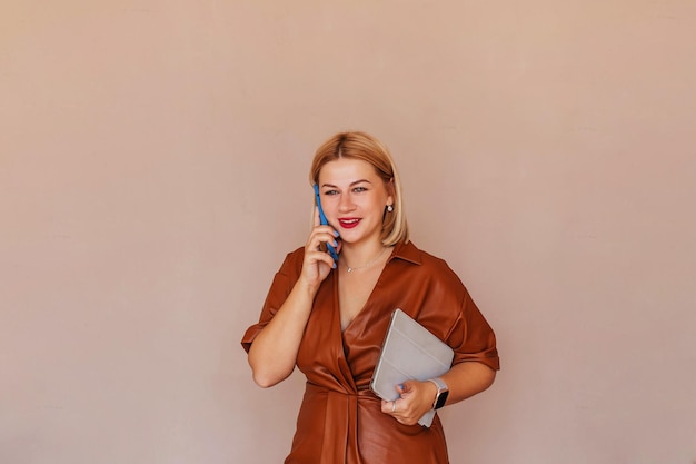 Young beautiful businesswoman in a brown dress talking on the phone with a laptop in her hands Young