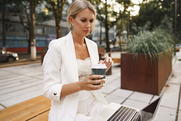 Young beautiful businesswoman working on laptop sitting on the bench in the street