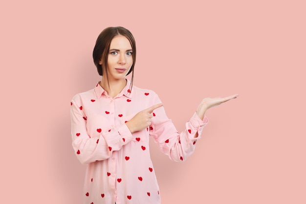 Young beautiful calm girl on a pink background in a pink shirt with red hearts points his fingers at the place for promotions offers or advertising