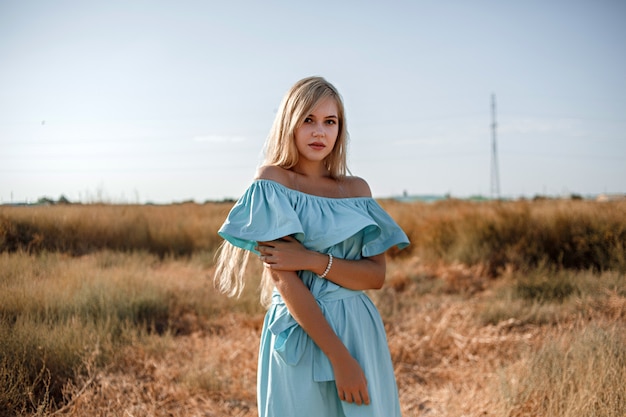 Young beautiful caucasian blonde girl in light blue dress stands on the field with the sun-scorched grass