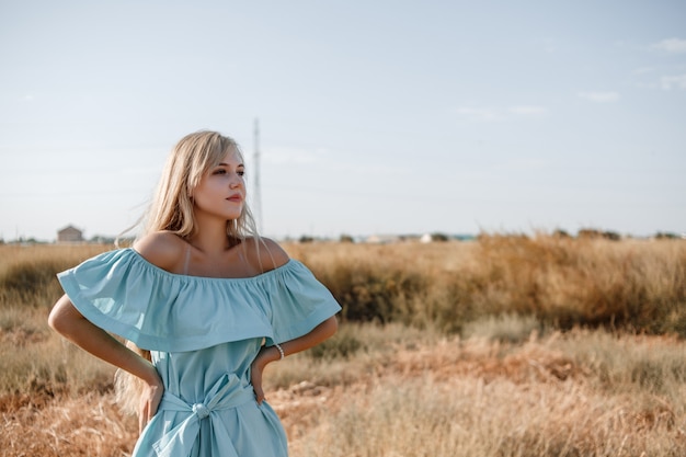 Young beautiful caucasian blonde girl in light blue dress stands on the field with the sun-scorched grass