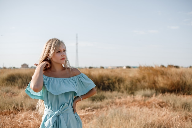 Young beautiful caucasian blonde girl in light blue dress stands on the field with the sun-scorched grass