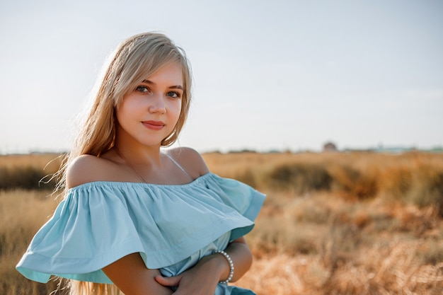 Young beautiful caucasian blonde girl in light blue dress stands on the field with the sun-scorched grass