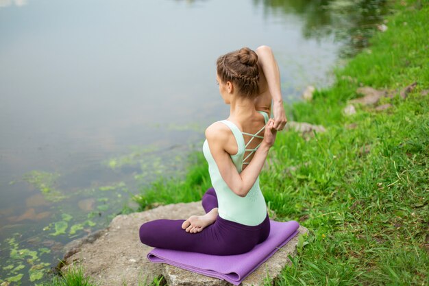 Young beautiful caucasian brunette girl doing yoga on a green lawn against the background of the river