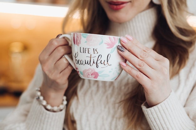 Young beautiful caucasian female with long chestnut hair in white sweater holds a white cup of coffe or tea, drinks it and enjoys of her life