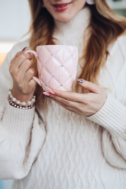 Young beautiful caucasian female with long chestnut hair in white sweater holds a white cup of coffe or tea and drinks it