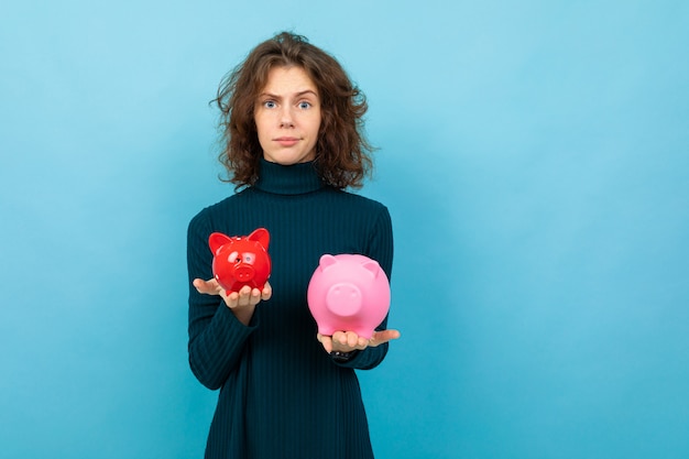 Young and beautiful caucasian girl with curly hair keeps small red pig moneybox, big pink pig moneybox and smiles, portrait isolated on blue background