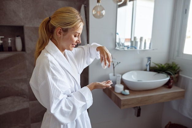 Young beautiful caucasian woman pouring body lotion on hand in bathroom antiaging concept